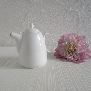 Könnte beinhalten: Eine glänzende, weiße Keramik-Teekanne mit gebogenem Griff und kleinem Deckel. Eine große, hellviolette Dahlienblüte liegt neben der Teekanne. Die Teekanne und die Blume stehen auf einer weißen Oberfläche mit strukturiertem, weißem Hintergrund.