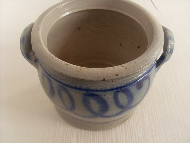 May include: A white ceramic crock with two handles and a blue and white design. The crock has a lid and is decorated with a repeating pattern of blue circles.