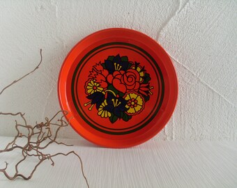 May include: A round orange metal tray with a floral design in black, yellow, red, and blue. The tray has a raised rim and a smooth surface.