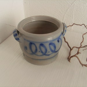 May include: A gray stoneware crock with blue painted designs and two handles. The crock has a lid and is decorated with a blue painted design of three circles.