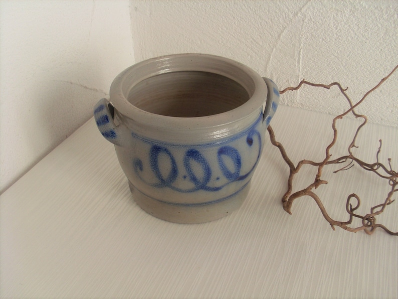 May include: A gray stoneware crock with blue painted designs and two handles. The crock has a lid and is decorated with a blue painted design of three circles.