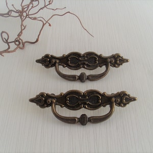 Könnte beinhalten: Zwei verzierte Messing-Schrankgriffe mit einem dekorativen Design. Die Griffe haben eine geschwungene Form und einen zentralen Ring.