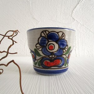 Könnte beinhalten: Eine weiße Keramiktasse mit einem blauen und roten Blumendesign. Die Tasse hat einen breiten Rand und einen abgerundeten Boden.