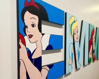 Könnte beinhalten: Holzbuchstaben, die mit Disney-Prinzessinnen bemalt sind und den Namen "EMMA" buchstabieren. Der Buchstabe "E" zeigt Schneewittchen, das einen roten Apfel hält, der Buchstabe "M" zeigt Aschenputtel in einem blauen Kleid und der Buchstabe "A" zeigt Arielle in einem grünen Kleid.