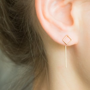 May include: Gold earrings with a square shape and a long, thin bar hanging down.