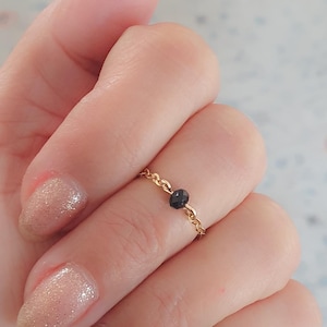 Può includere: Un delicato anello a catena dorato con una singola pietra preziosa nera. L'anello è indossato su un dito con uno smalto per unghie rosa chiaro con glitter.