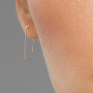 May include: A close-up of a person's face with a gold earring in the shape of a rectangle. The earring is simple and minimalist.