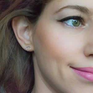 Pu&ograve; includere: Primo piano del viso di una donna con un piccolo orecchino esagonale d'argento nell'orecchio sinistro. Il suo trucco &egrave; naturale con un tocco di fard e i suoi occhi sono evidenziati da eyeliner e mascara. Sorride leggermente.