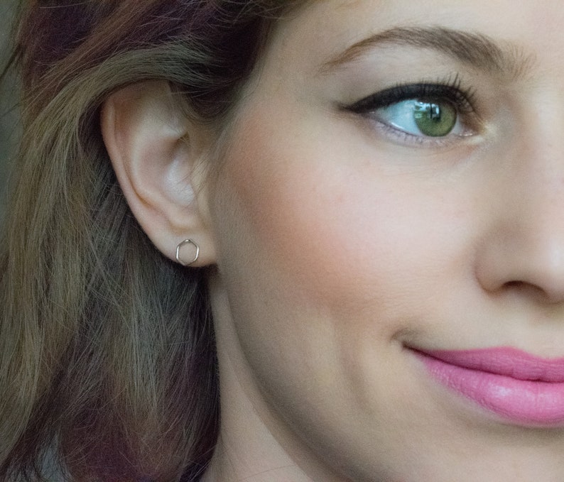 Pu&ograve; includere: Primo piano del viso di una donna con un piccolo orecchino esagonale d'argento nell'orecchio sinistro. Il suo trucco &egrave; naturale con un tocco di fard e i suoi occhi sono evidenziati da eyeliner e mascara. Sorride leggermente.
