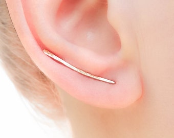 May include: A thin, gold-colored ear cuff with a slightly curved shape. The cuff is worn on the upper part of the ear.