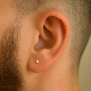 May include: A close-up of a person's ear with a small, round, gold stud earring. The earring is centered on the earlobe. The background is blurred, with the person's skin and hair visible.