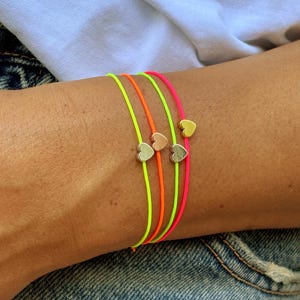 Könnte beinhalten: Drei dünne Kordelarmbänder in Neongelb, Neonorange und Neonpink. Jedes Armband ist mit einem kleinen, metallischen Herzanhänger versehen. Die Herzen sind in Silber-, Roségold- und Goldtönen gehalten und mittig auf den Kordeln platziert.