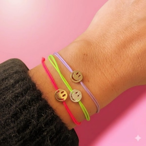 Könnte beinhalten: Drei Kordelarmbänder in Neonpink, Neongrün und Helllila, die jeweils mit einem kleinen, runden, goldfarbenen Metallanhänger mit ausgestanztem Smiley-Gesicht versehen sind. Die Armbänder sind dünn und verstellbar und werden zusammen getragen.