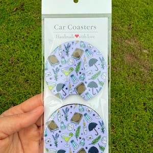 May include: Two circular car coasters in a clear plastic package with a header card labeled Car Coasters Handmade with love. The coasters feature a light purple background with a repeating pattern of books, umbrellas, candles, cocktails, and lavender.