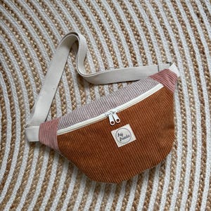Op de afbeelding: Een corduroy heuptas in bruin, roze en crèmekleur. De tas heeft een witte rits en een crèmekleurige verstelbare riem. Een klein wit label met de tekst "hej frieda" is op de voorkant genaaid.