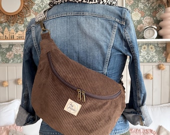 Peut inclure: Un sac bandoulière en velours côtelé marron avec une fermeture éclair incurvée et un écusson rectangulaire blanc portant le texte "hej frieda" en noir. Le sac comprend une sangle à motif chevron et des détails en laiton vieilli.