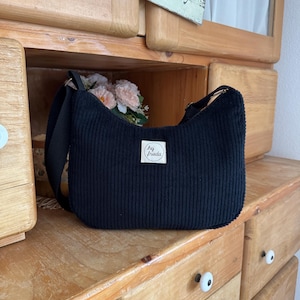 May include: Black corduroy shoulder bag with a rectangular shape and adjustable strap. The bag features a small white tag with the text "hej Frieda". The bag is displayed on a wooden surface.