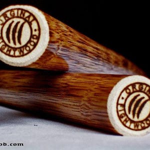 Könnte beinhalten: Zwei Holzstäbe mit hellbraunem Finish. Die Enden weisen ein kreisförmiges Design mit dem Text "ORIGINAL FIGHTWOOD" und einem stilisierten Logo auf. Die Stäbe sind abgewinkelt, wobei die Enden im Fokus stehen, und die Website Fightwood.com ist sichtbar.
