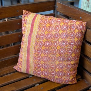 Peut inclure: Un coussin de décoration rouge, jaune et blanc avec un motif géométrique. Le coussin est posé sur un banc en bois.