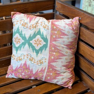 Peut inclure: Un coussin de décoration rose, vert et or avec un motif géométrique. Le coussin est posé sur un banc en bois.