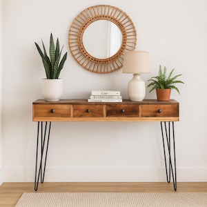 Può includere: Una consolle in legno con quattro cassetti e gambe a forcina nere. Uno specchio rotondo e intrecciato è appeso sopra il tavolo, decorato con una lampada, piante e libri. Il tavolo è in legno chiaro e scuro.