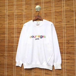 May include: White sweatshirt featuring the embroidered text "courr&egrave;ges paris" in rainbow hues. The crew neck sweatshirt has long sleeves. It is displayed on a wooden hanger against a bamboo backdrop.