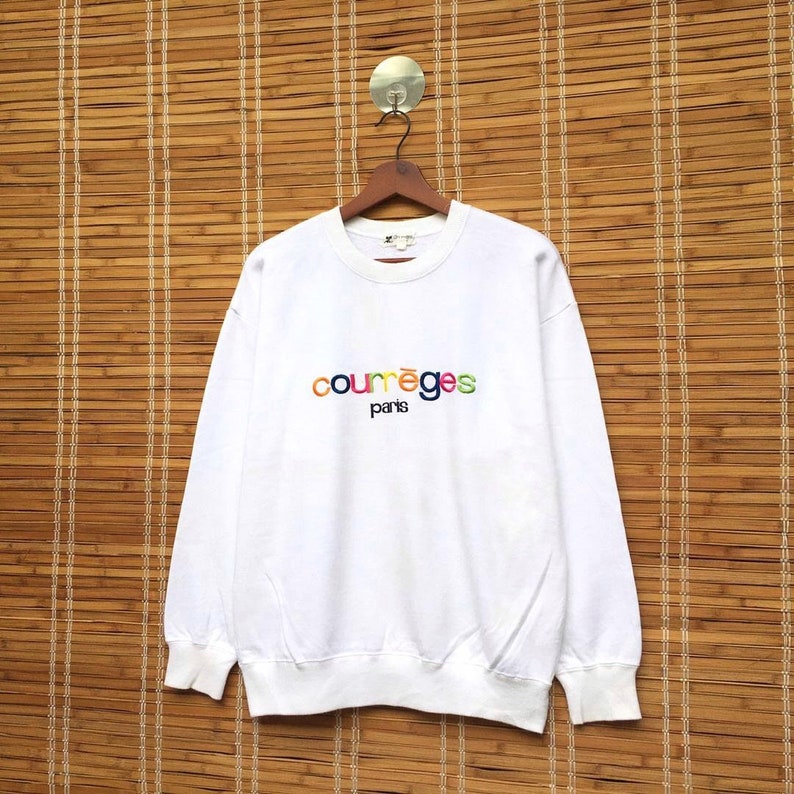 May include: White sweatshirt featuring the embroidered text "courr&egrave;ges paris" in rainbow hues. The crew neck sweatshirt has long sleeves. It is displayed on a wooden hanger against a bamboo backdrop.