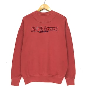 May include: Red sweatshirt with a crew neck and long sleeves. The sweatshirt has the text "Ralph Lauren Chaps" embroidered on the front in red and black.