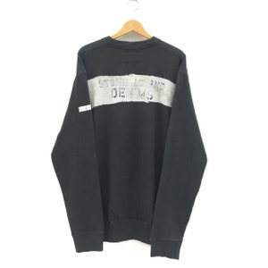 May include: Black sweatshirt with a wide gray stripe across the chest. The text "STONE ISLAND DENIMS" is printed in gray on the stripe. The sweatshirt is hanging on a wooden hanger.