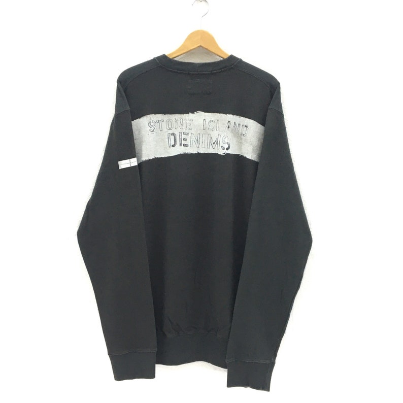 May include: Black sweatshirt with a wide gray stripe across the chest. The text "STONE ISLAND DENIMS" is printed in gray on the stripe. The sweatshirt is hanging on a wooden hanger.