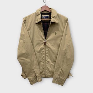 Puede incluir: Chaqueta beige con cremallera y cuello. La chaqueta presenta un pequeño logotipo bordado en el pecho y un forro a cuadros visible en el cuello. La chaqueta está colgada de una percha de madera.