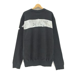 以下が含まれることがあります： 黒色の長袖スウェットシャツ。胸には、かすれたグレーの水平ストライプが入っています。「STONE ISLAND DENIMS」の文字が、ストライプの中に色あせた質感でプリントされています。スウェットシャツは木製のハンガーに掛けられています。