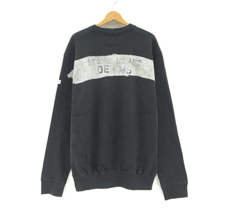 May include: Black sweatshirt with long sleeves, featuring a distressed gray horizontal stripe across the chest. The text "STONE ISLAND DENIMS" is printed in a faded, textured style within the stripe. The sweatshirt is hanging on a wooden hanger.