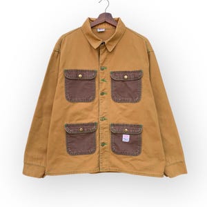 May include: A tan work jacket with four front pockets and a button-up closure. The pockets are a darker brown color with green stitching. The jacket is hanging on a wooden hanger.