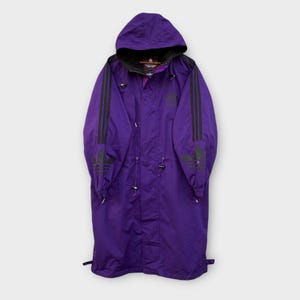 May include: A long purple hooded jacket with black Adidas logo stripes on the sleeves and a black zipper. The jacket has a drawstring waist and a hood with a drawstring.