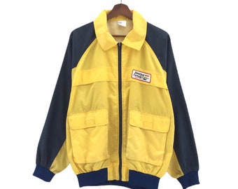 May include: A yellow and navy blue windbreaker jacket with a zipper closure and two large pockets. The jacket has a "Benson & Hedges Formula 1" logo on the right chest.