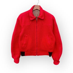 May include: A bright red jacket with a textured surface hangs from a wooden hanger. The jacket features a collar, a zipper closure, and two front pockets. Black trim accents the cuffs and hem. The jacket's style is reminiscent of a bomber jacket.