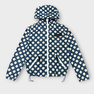 May include: A blue jacket with a white polka dot pattern. The jacket has a hood, a white zipper, and two front pockets. The jacket also has a drawstring at the bottom and a small patch on the upper right chest.