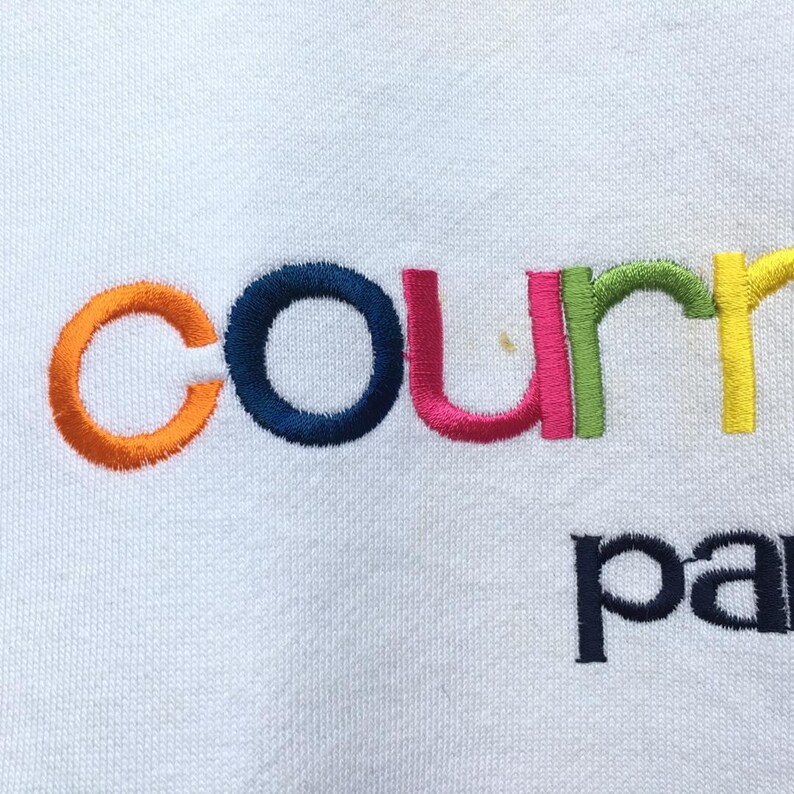 May include: White fabric with the embroidered word "court" in orange, navy blue, pink, green, and yellow. Below, the word "pa" is embroidered in navy blue. The fabric has a textured appearance.