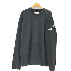 May include: A black crew neck sweatshirt with a small white and gray patch on the left sleeve. The sweatshirt is hanging on a wooden hanger against a white background. The brand name is visible on the tag.