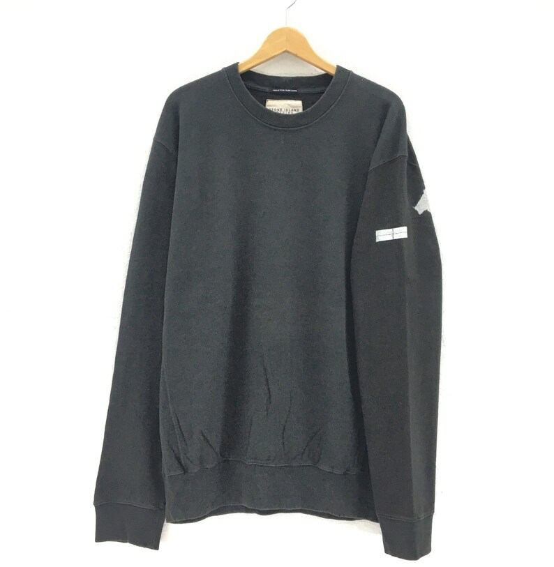 May include: A black crew neck sweatshirt with a small white and gray patch on the left sleeve. The sweatshirt is hanging on a wooden hanger against a white background. The brand name is visible on the tag.