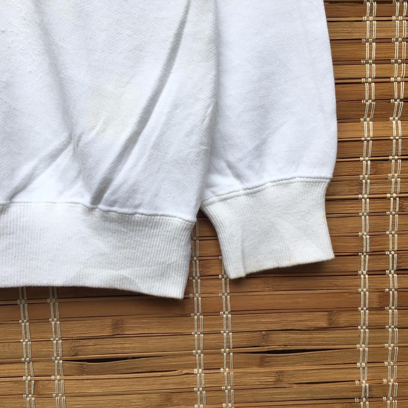 May include: A close-up view of a white sweatshirt, focusing on the ribbed hem and cuff. The fabric shows signs of wear, with some discolouration visible. The image emphasizes the garment's texture and design.
