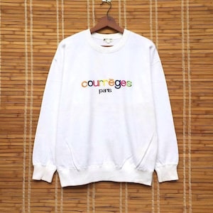 May include: White sweatshirt featuring the embroidered text "courr&egrave;ges paris" in rainbow colours. The long-sleeved top is displayed on a wooden hanger against a bamboo blind background.