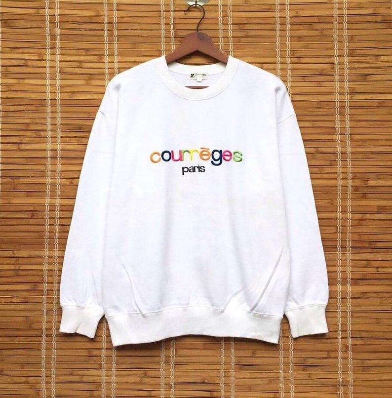 May include: White sweatshirt featuring the embroidered text "courr&egrave;ges paris" in rainbow colours. The long-sleeved top is displayed on a wooden hanger against a bamboo blind background.