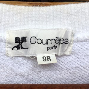 May include: A close-up shot of a white label with the black text "Courr&egrave;ges paris" and a logo. Below is a smaller label with the number "9R". The labels are attached to a white fabric. The fabric appears to be a sweater.