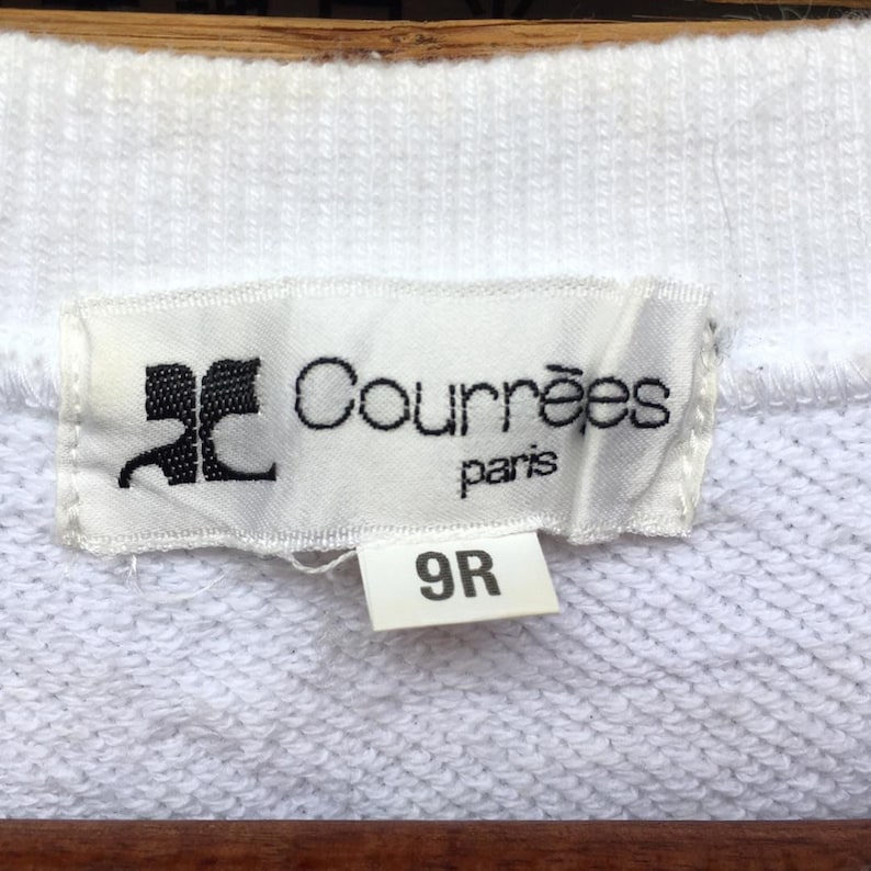 May include: A close-up shot of a white label with the black text "Courr&egrave;ges paris" and a logo. Below is a smaller label with the number "9R". The labels are attached to a white fabric. The fabric appears to be a sweater.