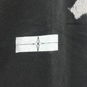 May include: Close-up of a black t-shirt with a white rectangular patch. The patch features a compass rose design in the center. The fabric has a textured appearance.