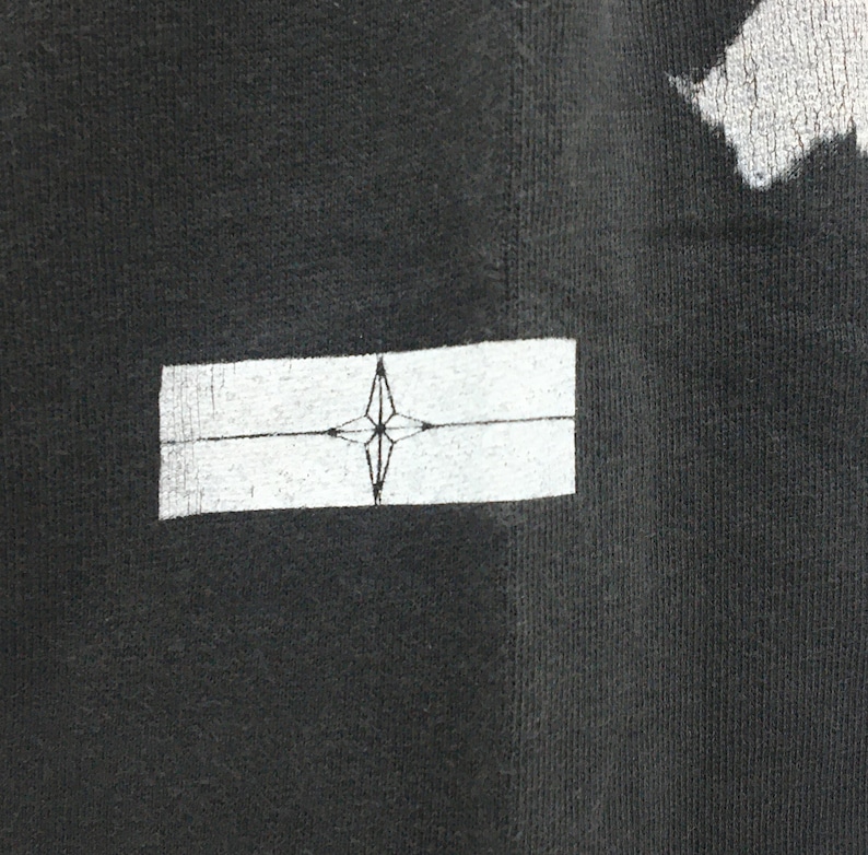 May include: Close-up of a black t-shirt with a white rectangular patch. The patch features a compass rose design in the center. The fabric has a textured appearance.