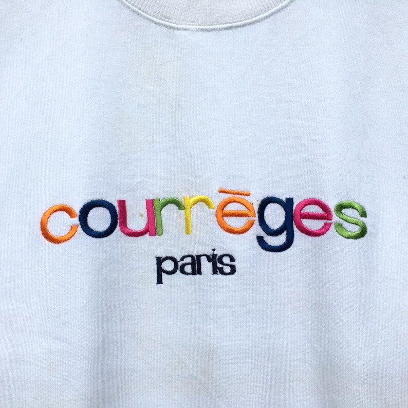 May include: White sweatshirt featuring the embroidered text "courr&egrave;ges paris". The brand name is in a rainbow of colours, with the city name in a smaller, dark blue font. A classic, casual garment.