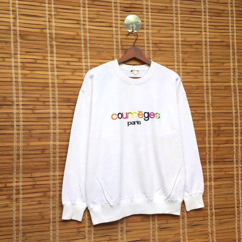 May include: White sweatshirt featuring the embroidered text "courr&egrave;ges paris" in rainbow hues. The long-sleeved top is displayed on a wooden hanger, set against a bamboo blind backdrop.